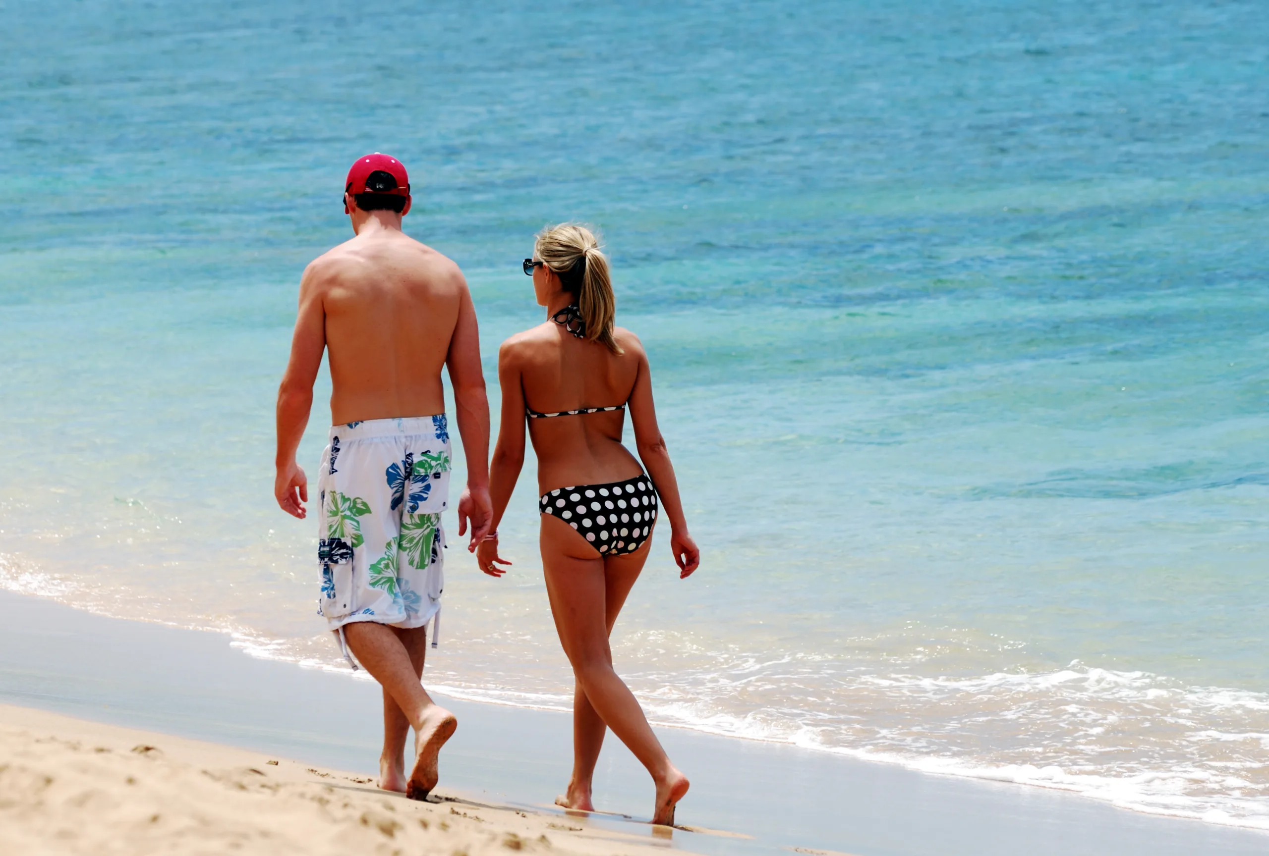 A honeymoon couple walks hand in hand along the shoreline of a Maui beach, barefoot in the sand as gentle waves meet the coast. The clear blue water and open shoreline create a relaxed, intimate setting that reflects the easy rhythm of a Hawaii honeymoon. Moments like these capture why Maui remains a favorite for couples seeking romance, warmth, and time together without distractions.