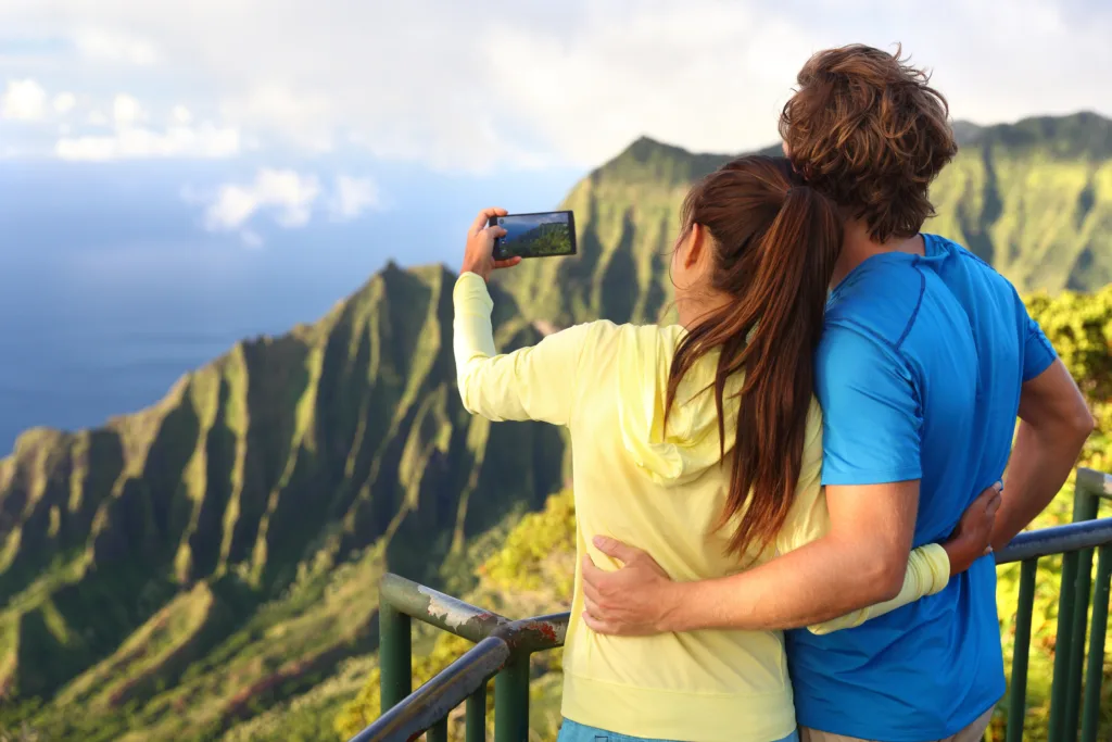 Honeymoon couple standing at a scenic Kauai lookout, taking a photo of the lush Na Pali Coast cliffs overlooking the ocean.