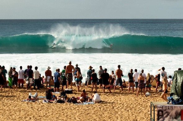 Spectators watch as surfers catch a huge wave barreling at Pipeline on Oahu's north shore