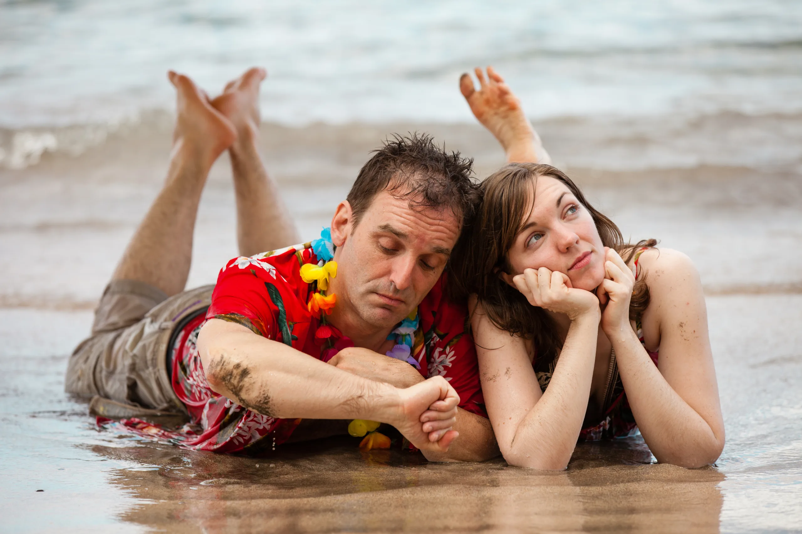 Couple lying on a sandy beach at the shoreline, relaxed and unhurried, embodying the laid-back pace of Hawaiian time.