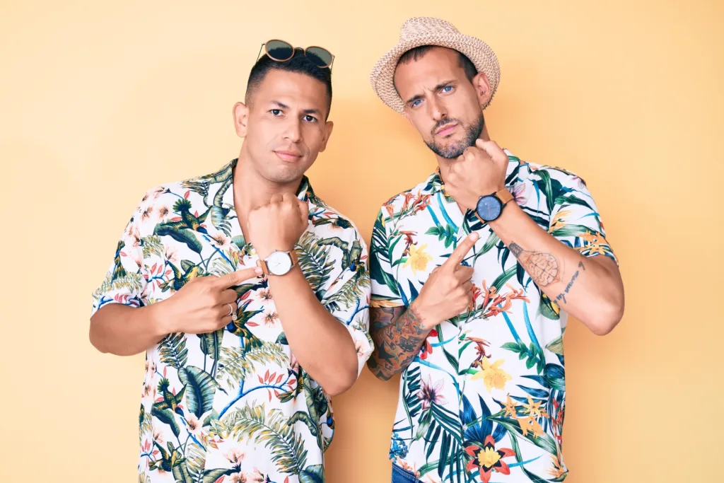 Two men wearing tropical shirts point to their wristwatches, highlighting time while dressed in relaxed island-style clothing.